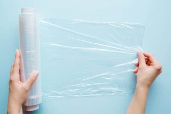 woman's hand using a roll of transparent polyethylene food film for packing products on the pastel blue table.