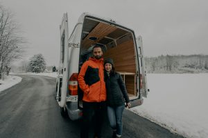 man in orange puffer jacket next to woman in black puffer jacket in front of a van stopped on a snowy road. Digital Nomads need storage