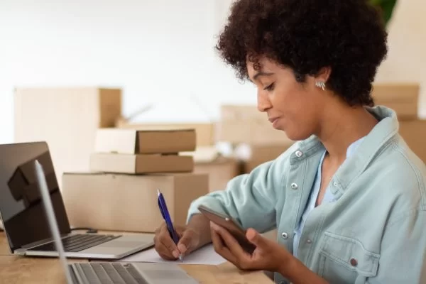 young startup owner holding smartphone checking order details attractive black woman working computer surrounded by cardboard parcels